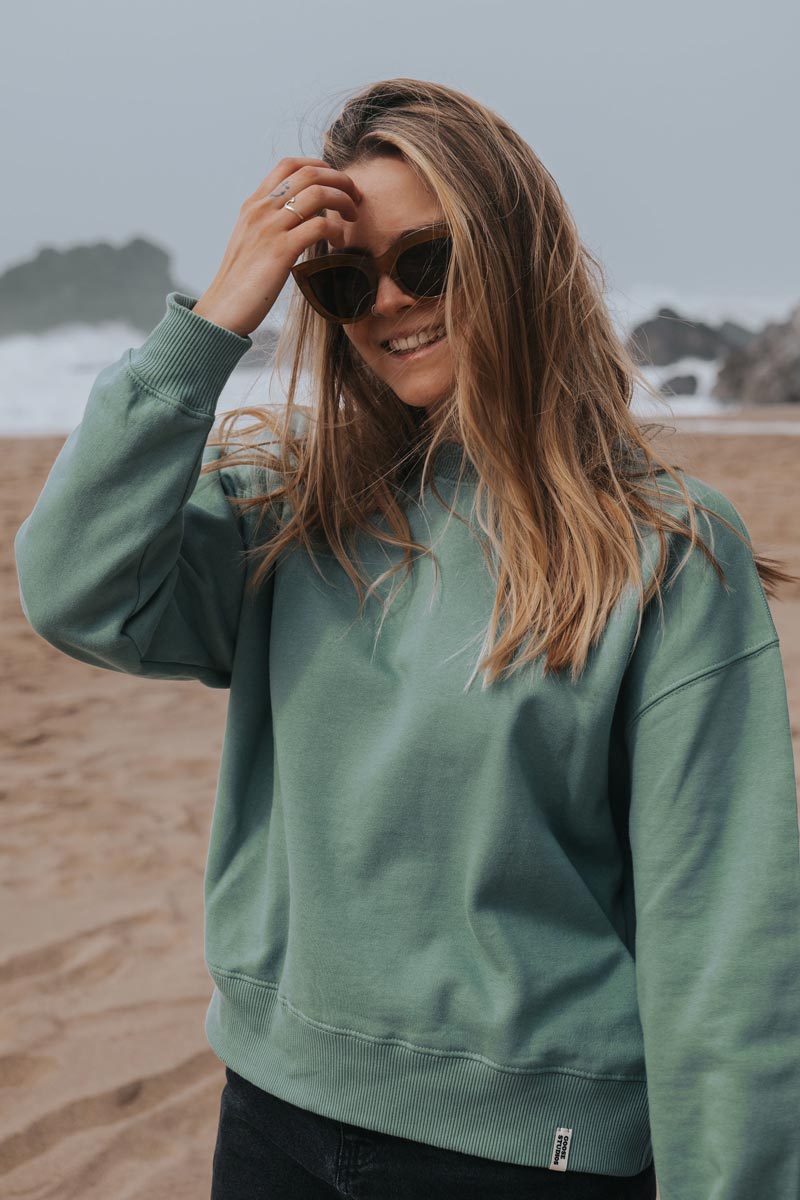 Woman wearing pastel green organic cotton sweatshirt from sustainable clothing brand Goose Studios whilst walking on the beach