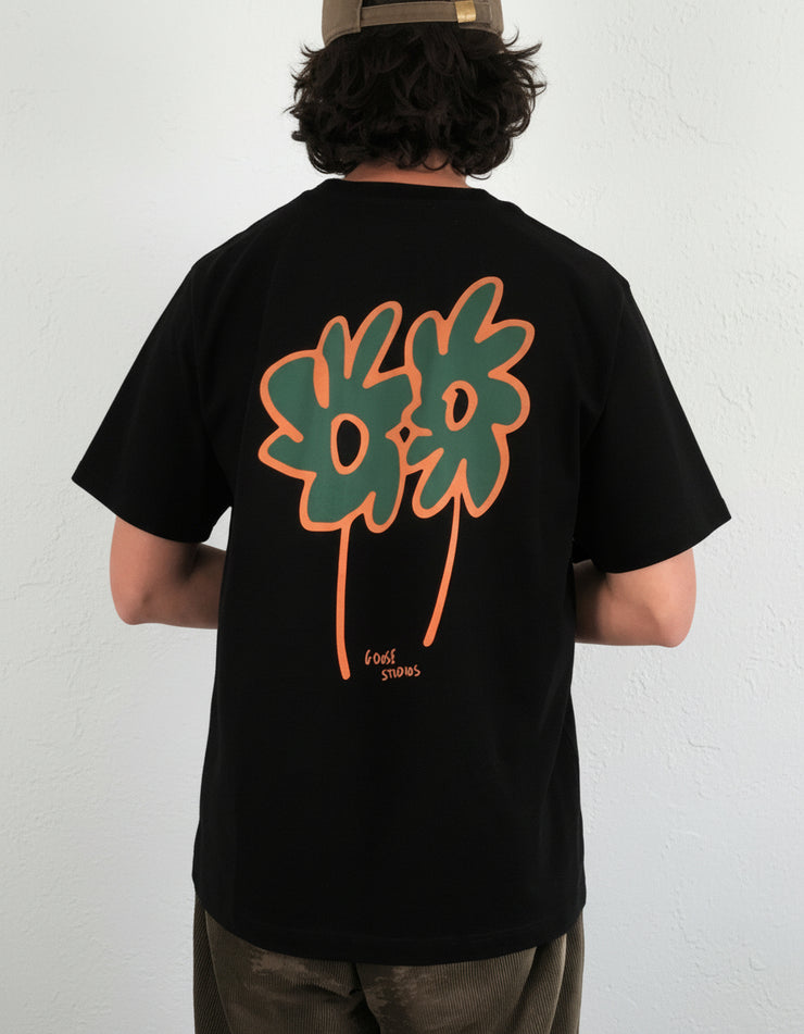 Man wearing a black heavyweight organic cotton t-shirt with a colourful flower design on the back, standing indoors.