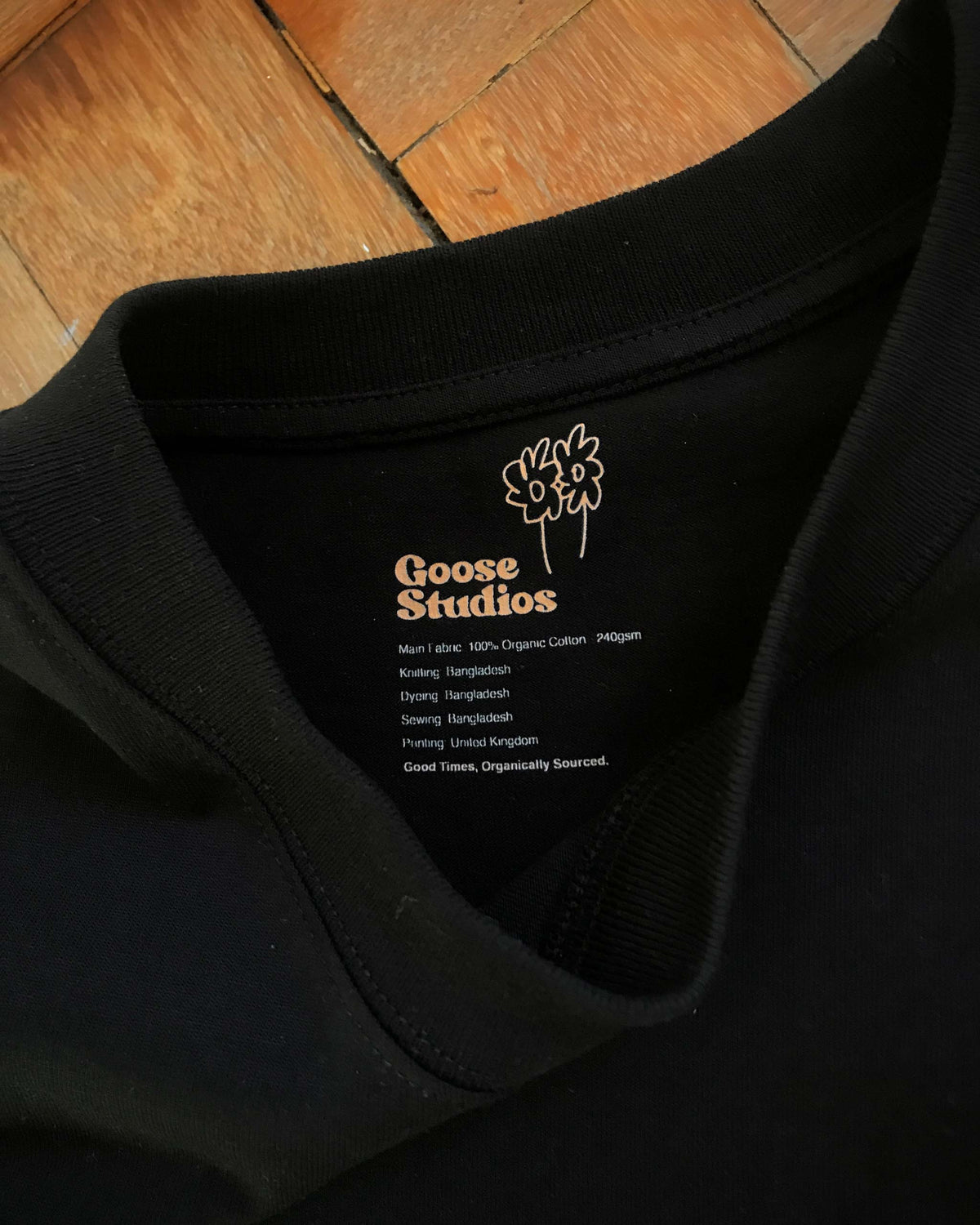 Black 240gsm relaxed fit men's t-shirt showing the collar branding with a 'Goose Studios' label, lying on a wooden floor