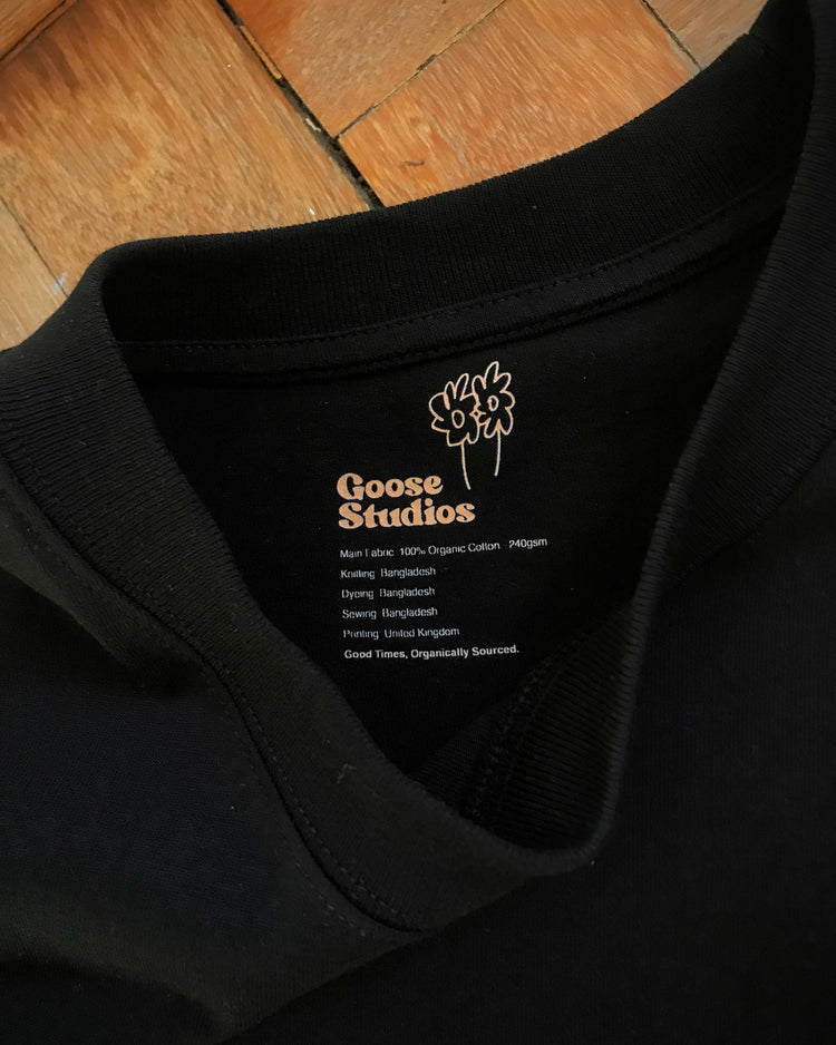Black 240gsm relaxed fit men's t-shirt showing the collar branding with a 'Goose Studios' label, lying on a wooden floor