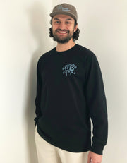 Man wearing a black long sleeve organic cotton t-shirt from Goose Studios with a logo and a cap, standing indoors.