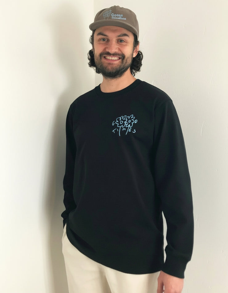 Man wearing a black long sleeve organic cotton t-shirt from Goose Studios with a logo and a cap, standing indoors.