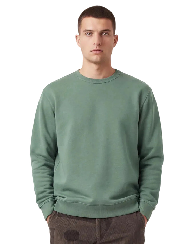 A man wearing a sage green organic cotton crewneck sweatshirt and dark brown pants, standing against a light studio background.