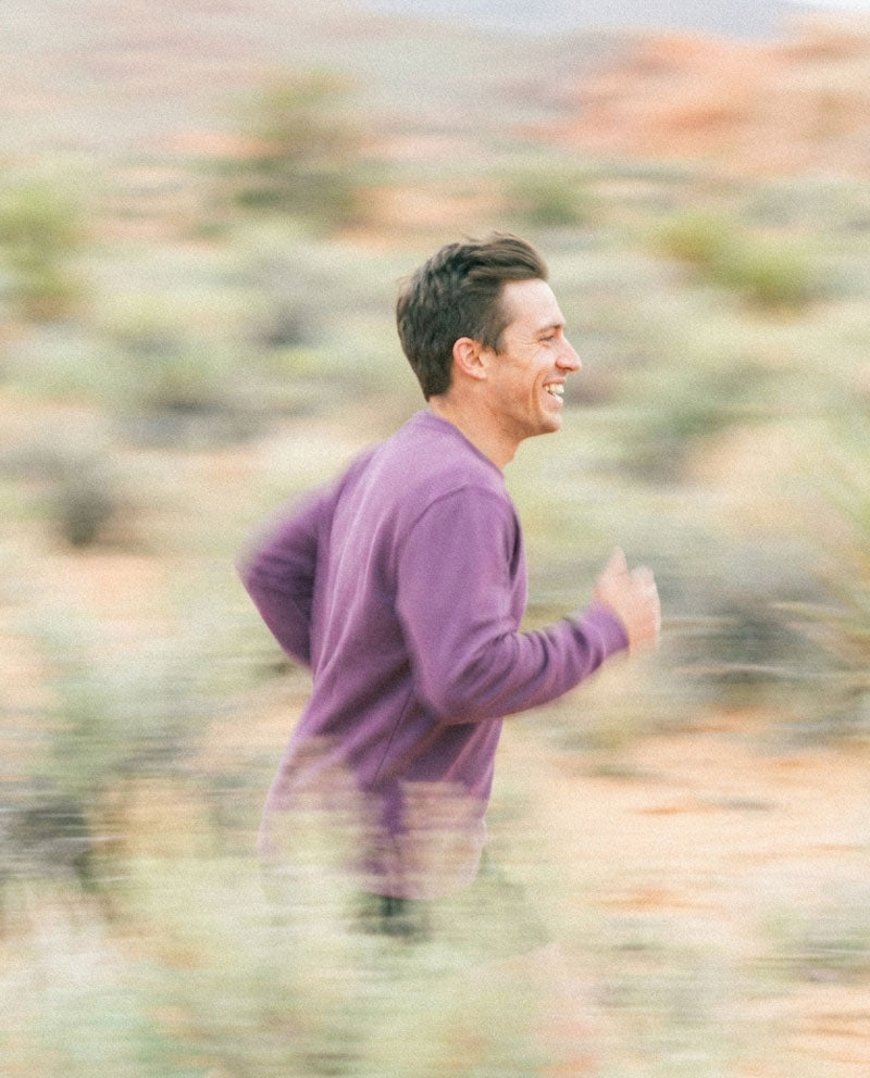A man wearing a purple long-sleeve organic sweatshirt is running through a blurred desert landscape with shrubs and reddish-brown hills in the background.
