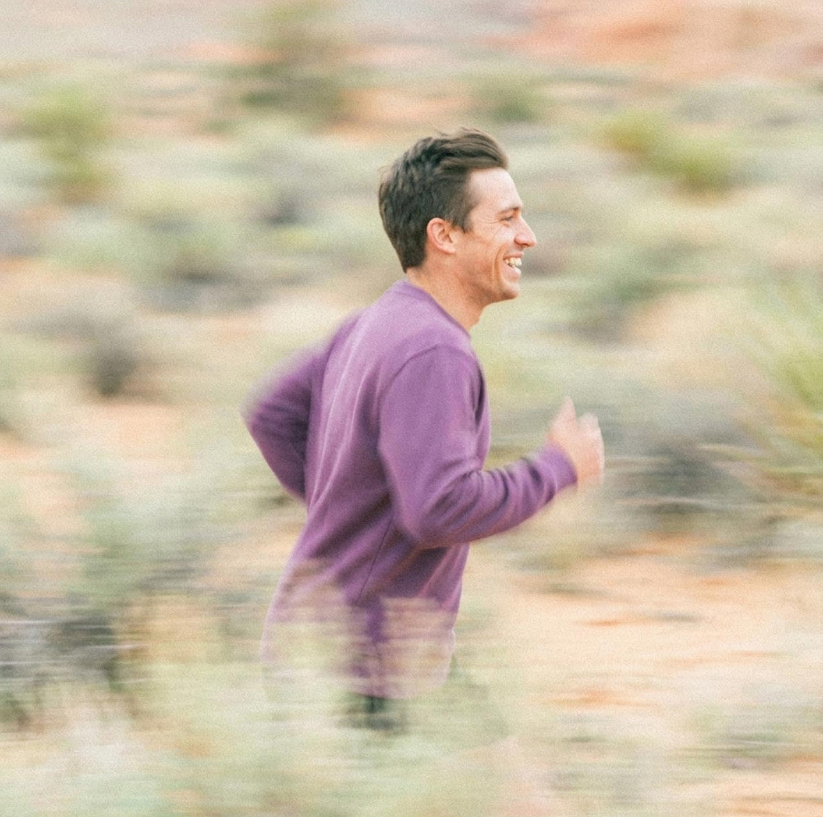 A man wearing a purple long-sleeve organic cotton sweatshirt is jogging outdoors in a blurred desert landscape.