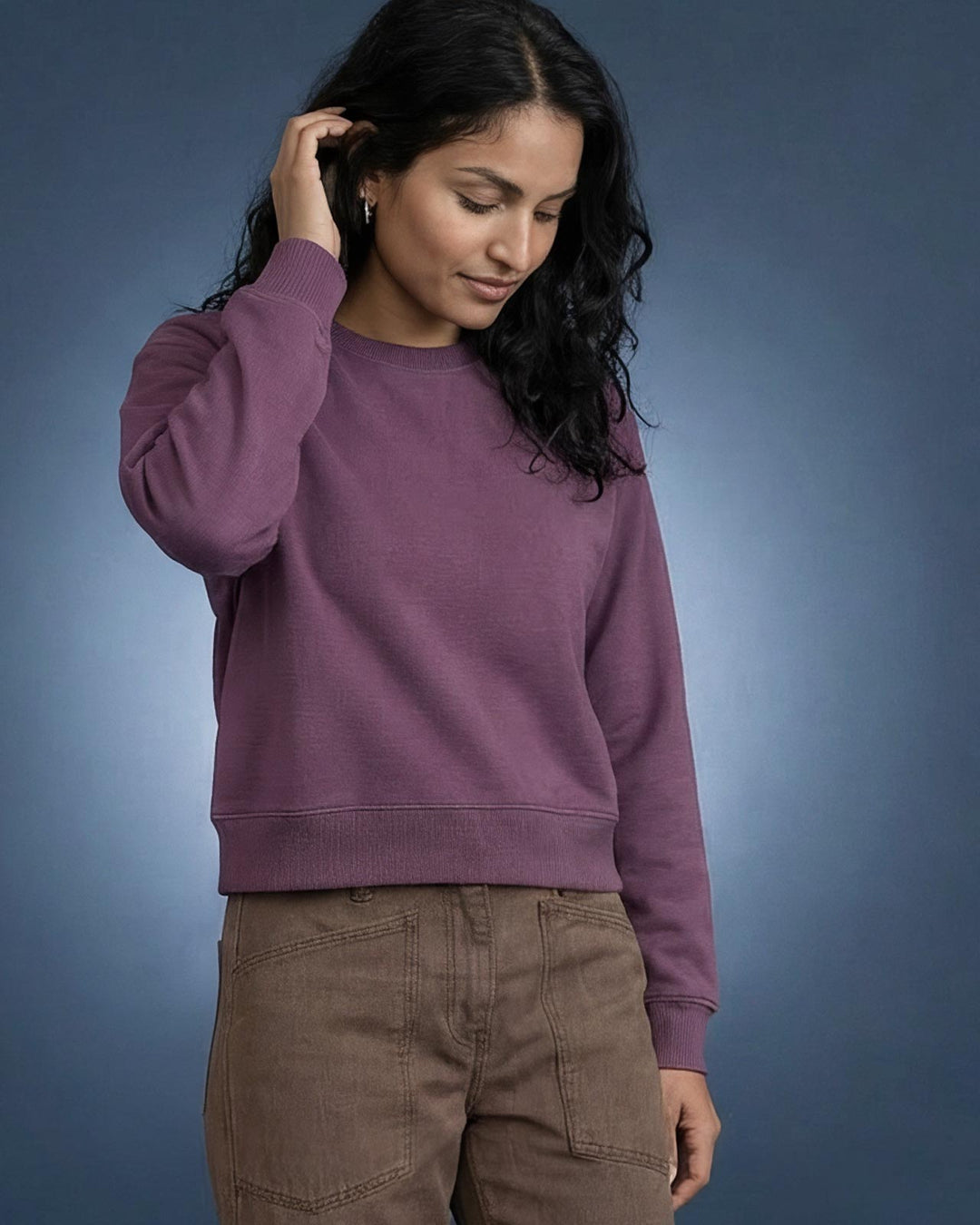 A woman wearing a berry purple crewneck organic sweatshirt and brown cargo pants, standing against a blue gradient studio background.