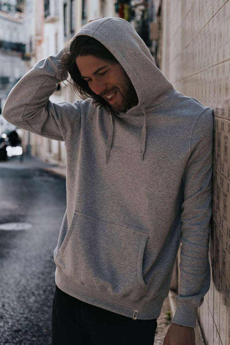 Man wearing a grey organic cotton hoodie with the hood up on the streets of Lisbon, hoodie features white label from Goose Studios an organic clothing brand from the UK